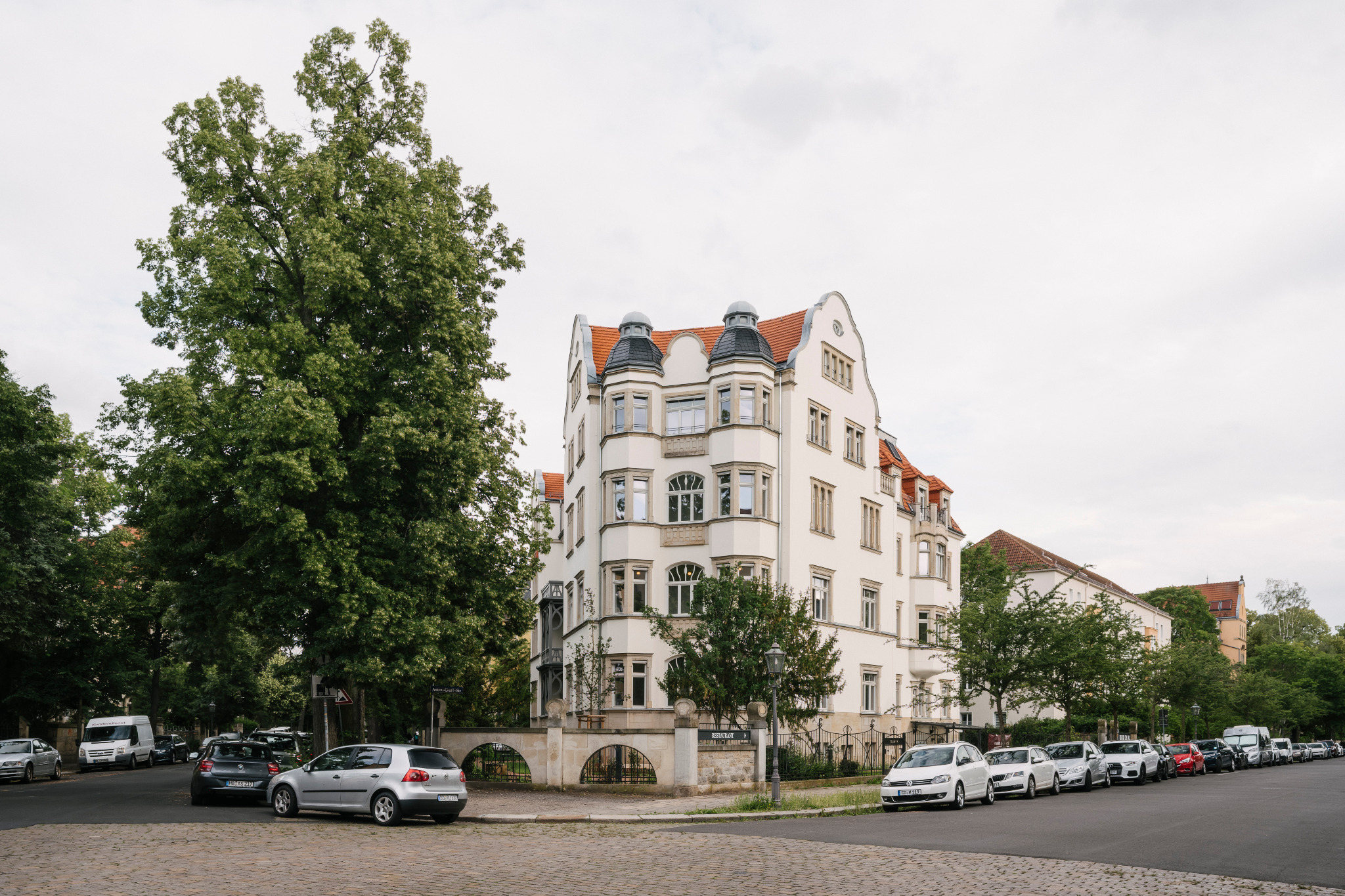 Gesamtansicht einer hellen Jugendstilvilla an einer Straßenecke mit großem Baumbestand.