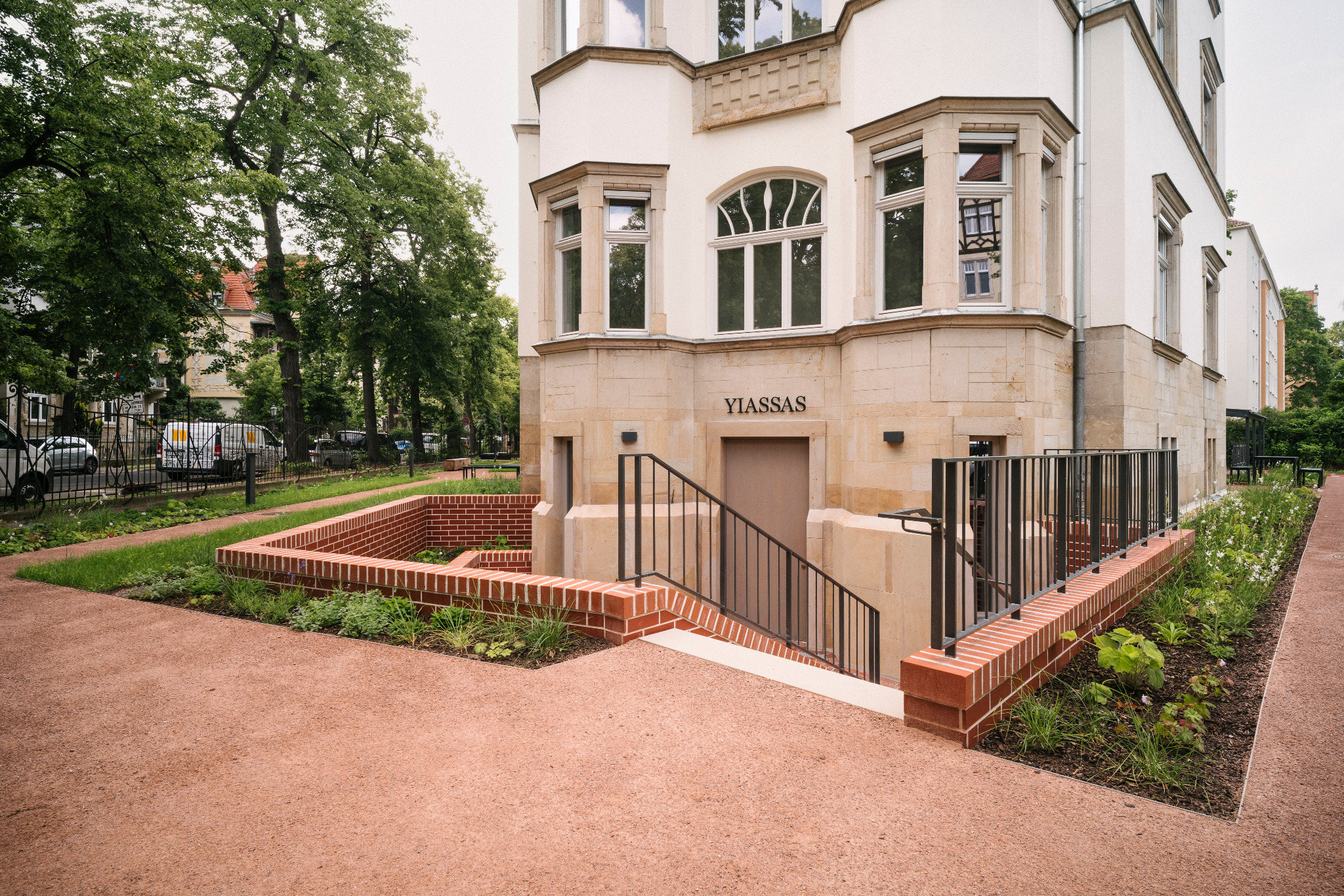 Seitlicher Gebäudeeingang mit Schriftzug YIASSAS, Treppen, roten Ziegelmauern und bepflanzten Außenanlagen.
