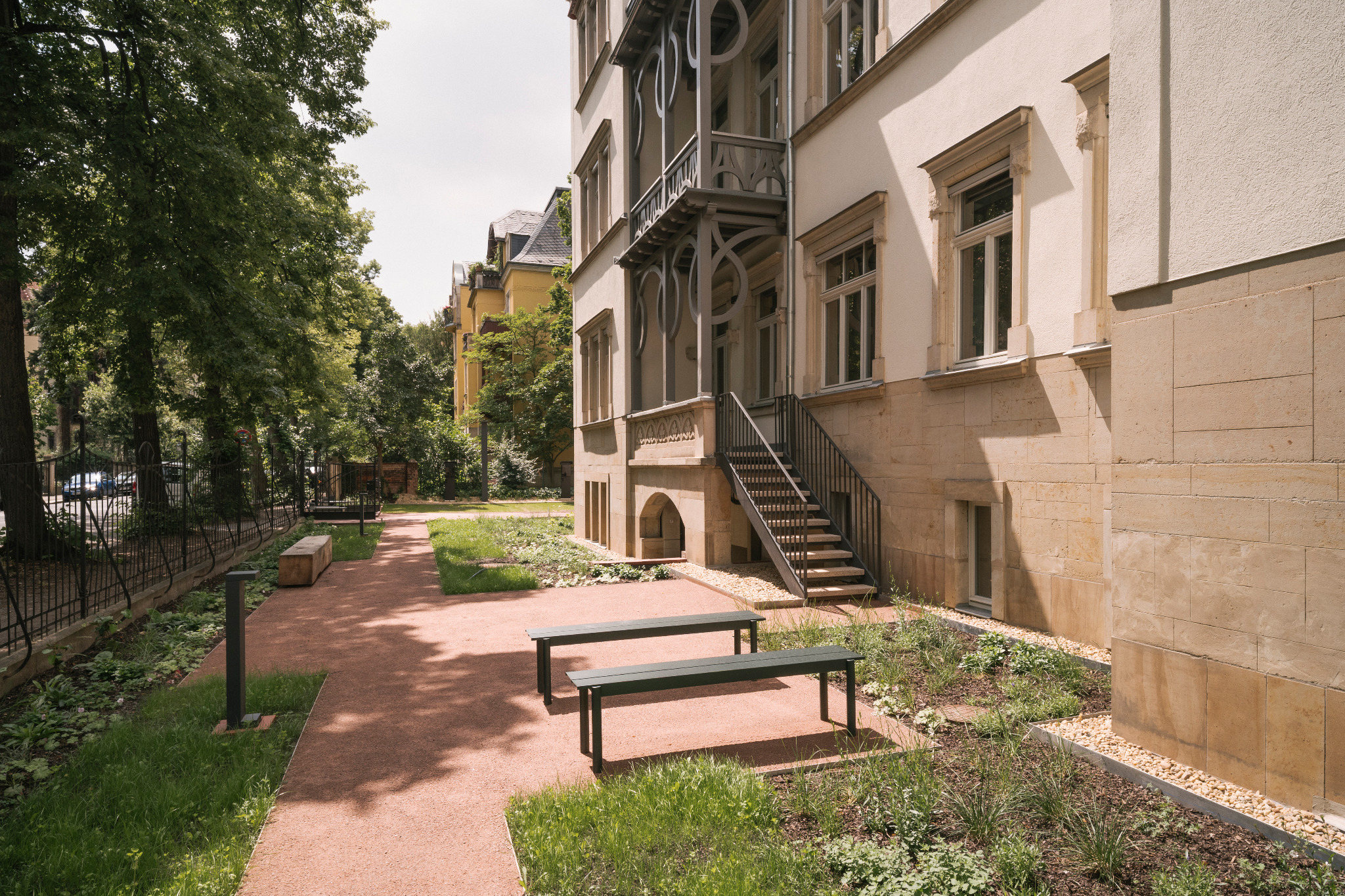 Seitliche Gartenansicht der Villa mit Wegen, Pflanzflächen, Sitzbänken und einer Außentreppe.