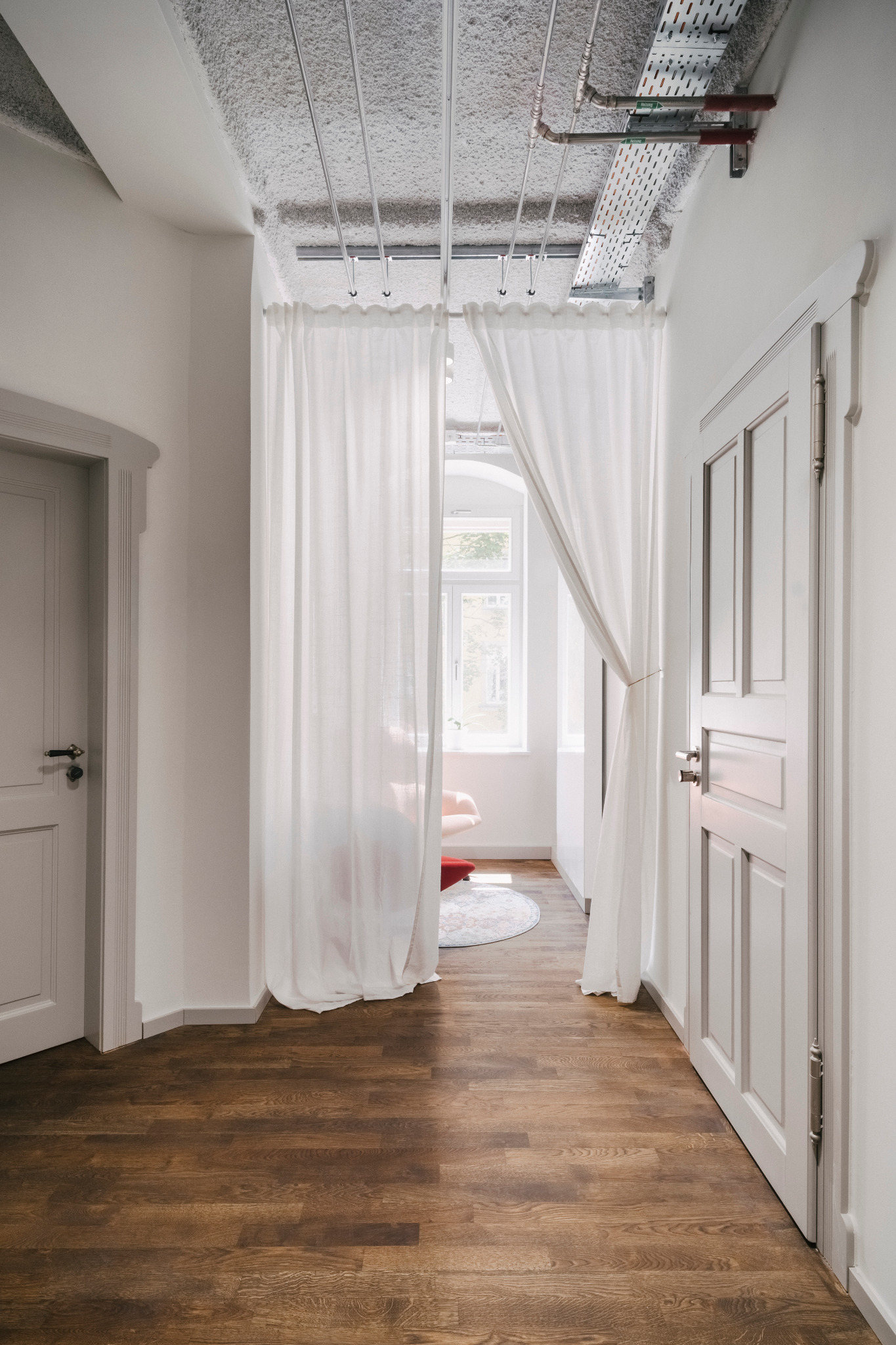 Heller Flur mit Holzboden, grauen Kassettentüren, weißen Vorhängen und sichtbaren Leitungen an der Decke.